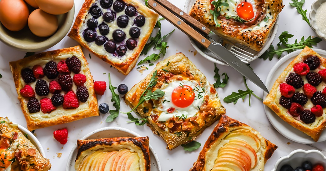 Overhead view of golden puff pastry tarts topped with berries, baked eggs, apples, and cream cheese, surrounded by fresh fruit and a bowl of eggs on a white table.